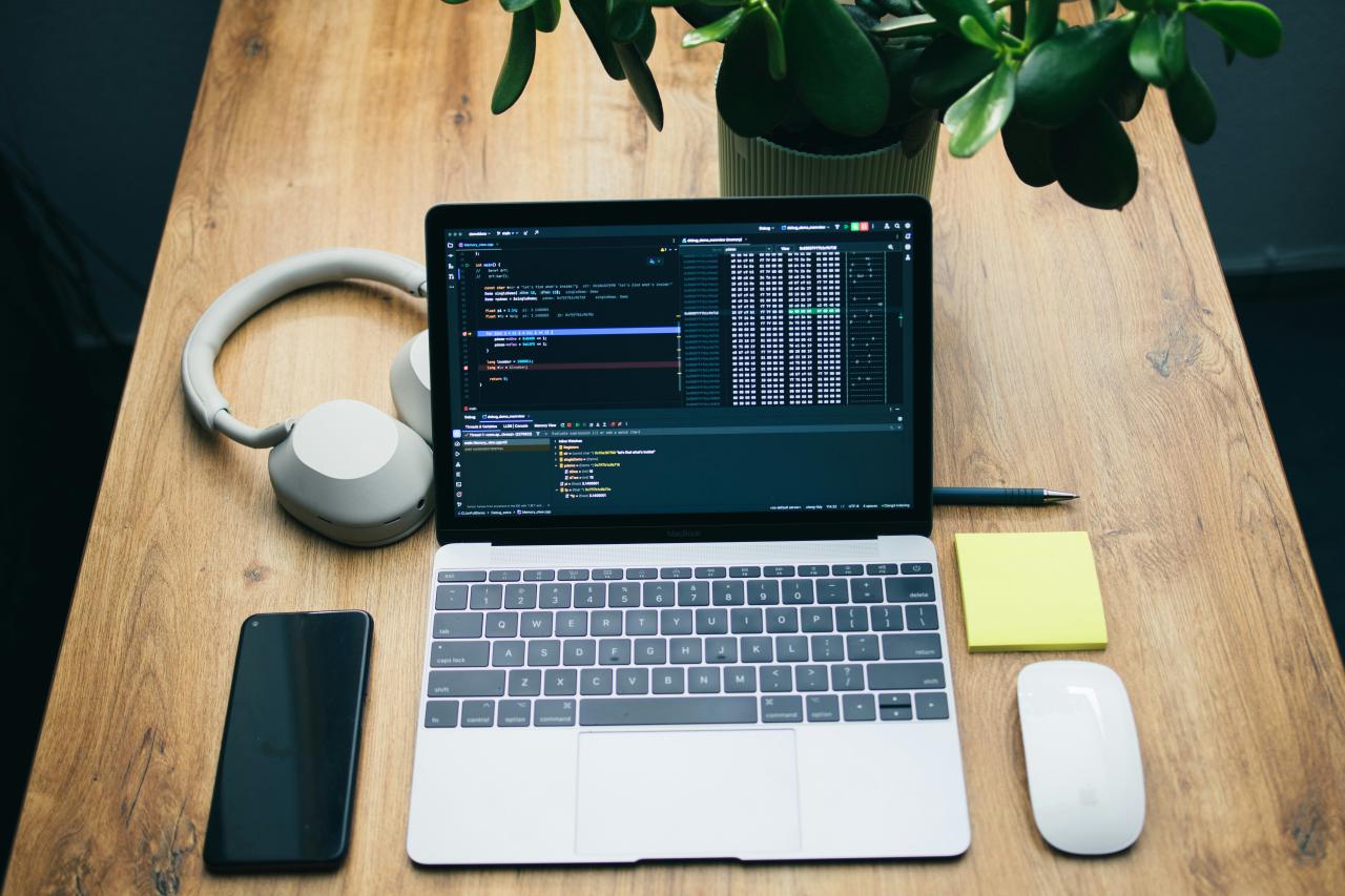 Laptop dengan kode, headphone, ponsel, dan mouse di atas meja.